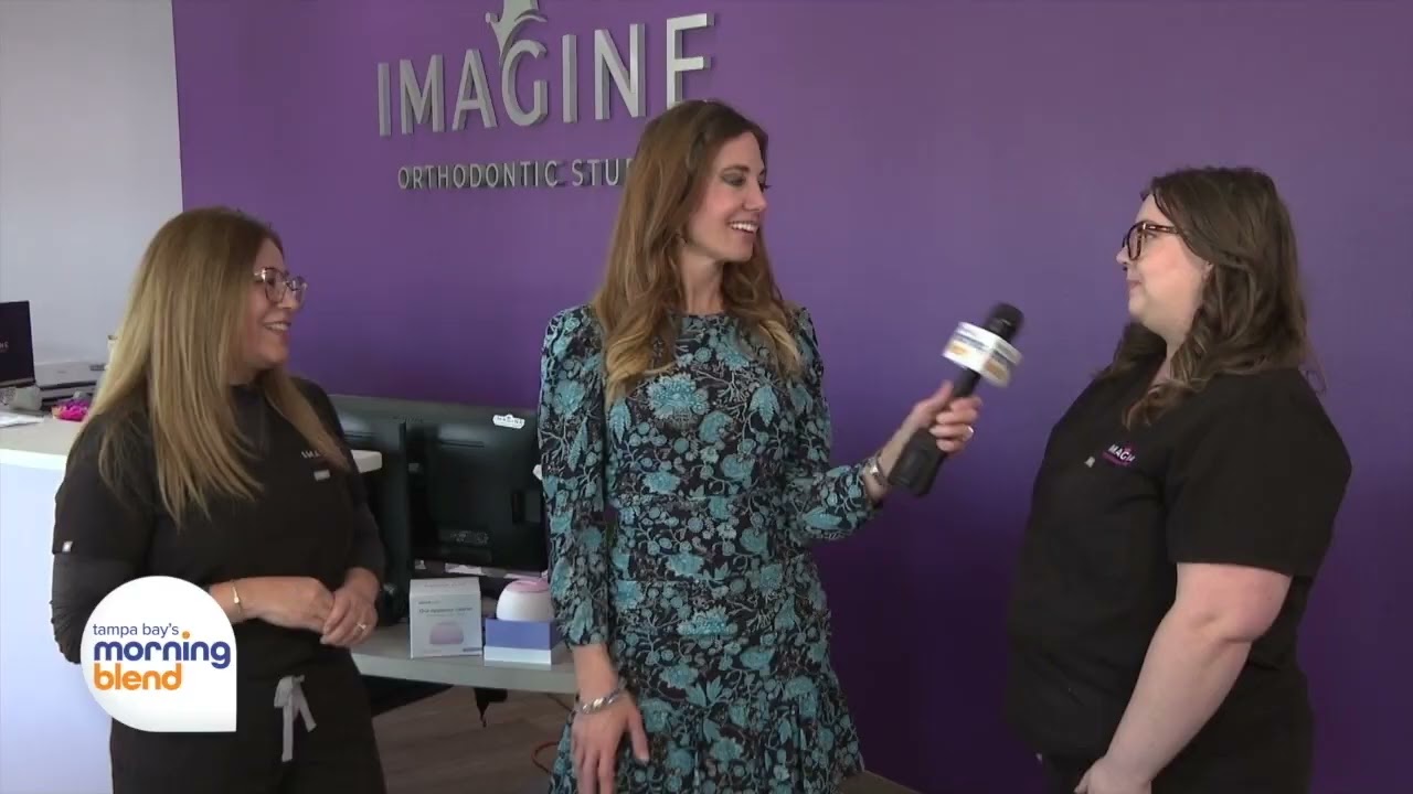 Three women interacting in Imagine Orthodontic Studio, featuring a purple wall with the studio's logo, showcasing a welcoming atmosphere for orthodontic consultations.